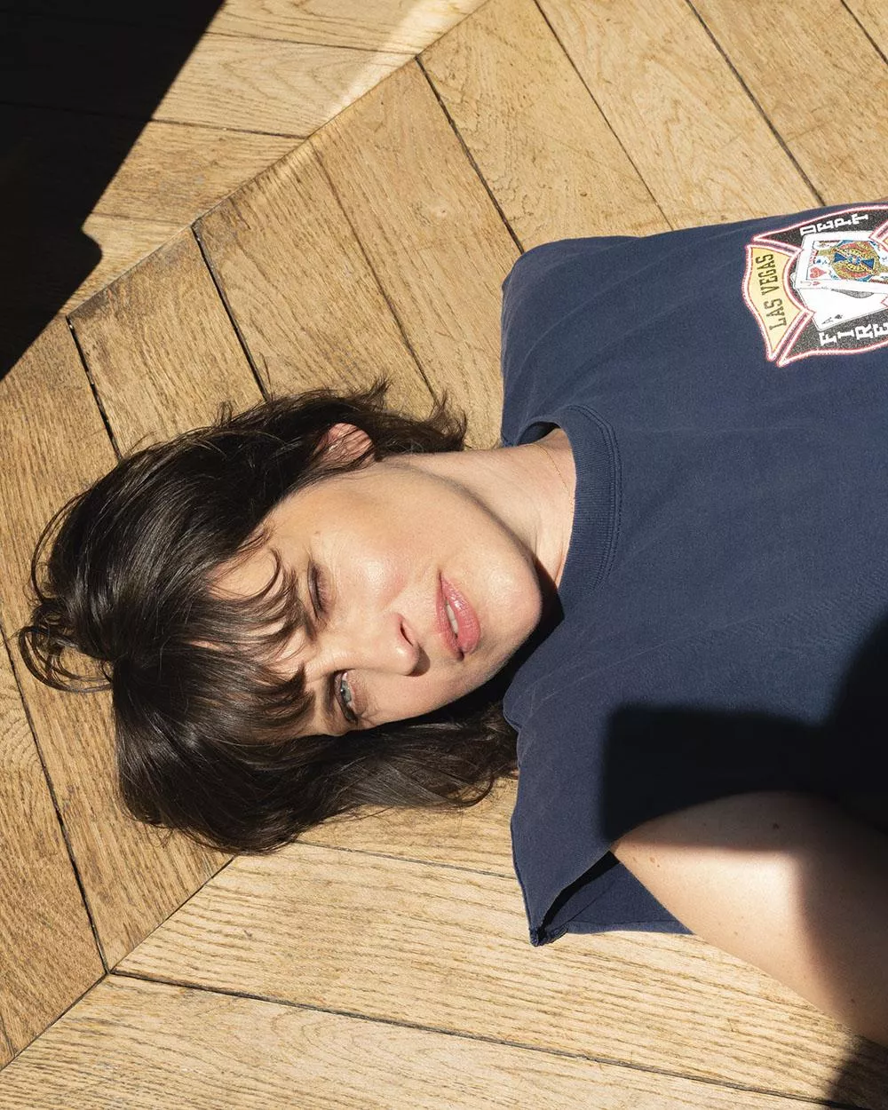Une personne est allongée sur un sol en bois, portant un t-shirt bleu avec un motif "Las Vegas". Elle est partiellement éclairée par le soleil.