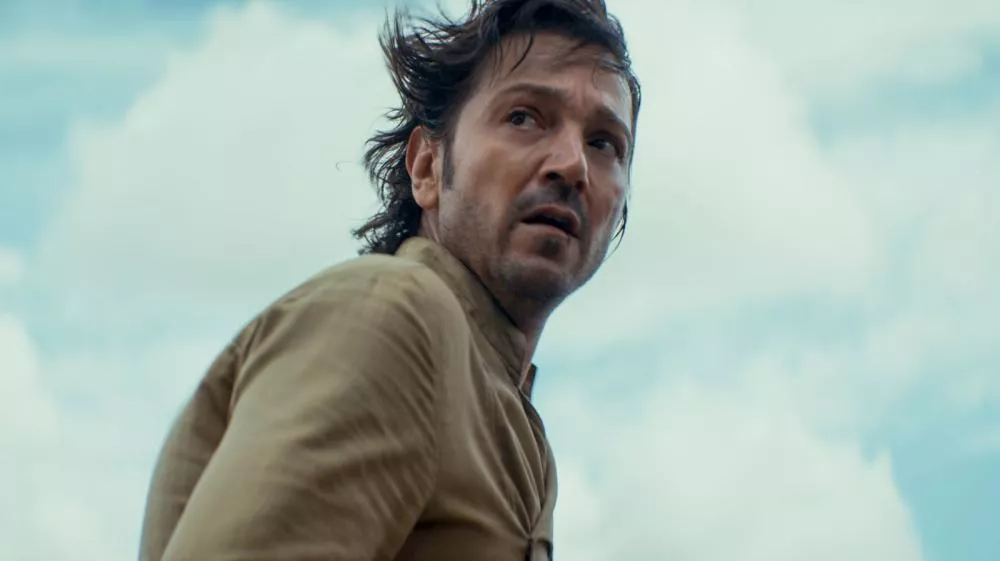 Un homme avec des cheveux ébouriffés regarde derrière lui avec une expression sérieuse, vêtu d'une chemise beige, sur un fond de ciel nuageux.
