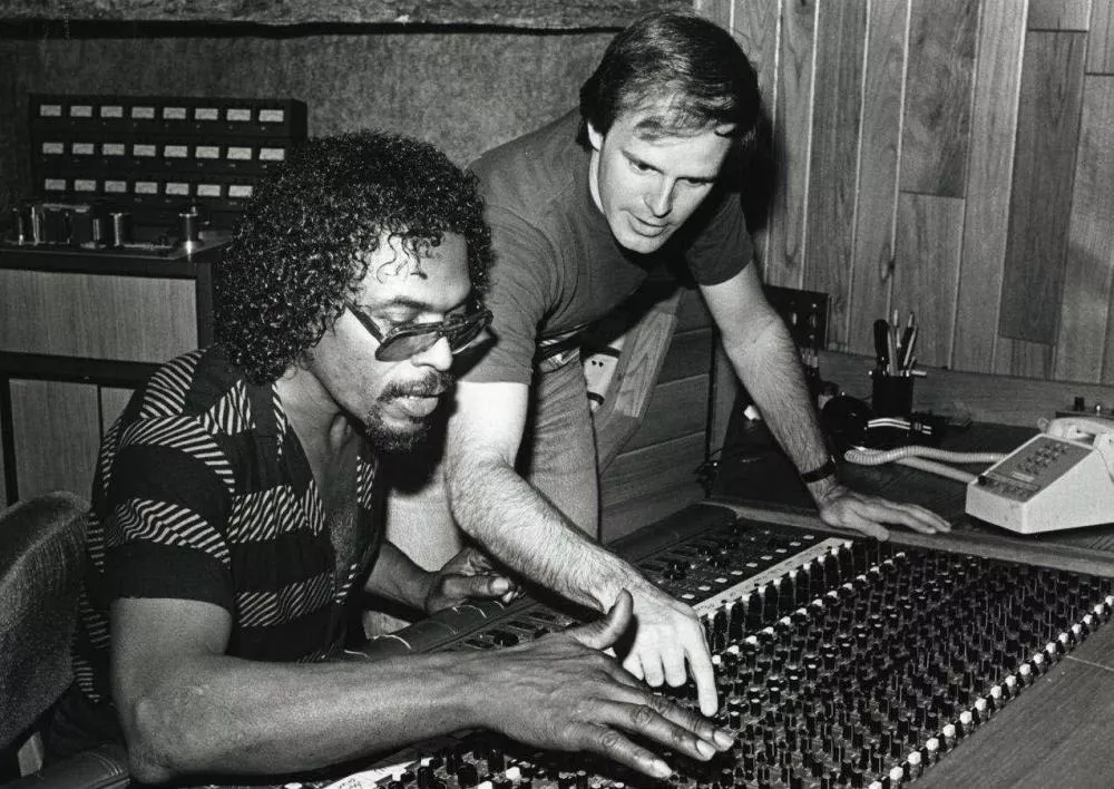 Deux hommes sont dans un studio d'enregistrement, concentrés sur une console de mixage. L'un porte des lunettes de soleil et une chemise rayée, l'autre est en t-shirt. Ils semblent collaborer sur des réglages audio.