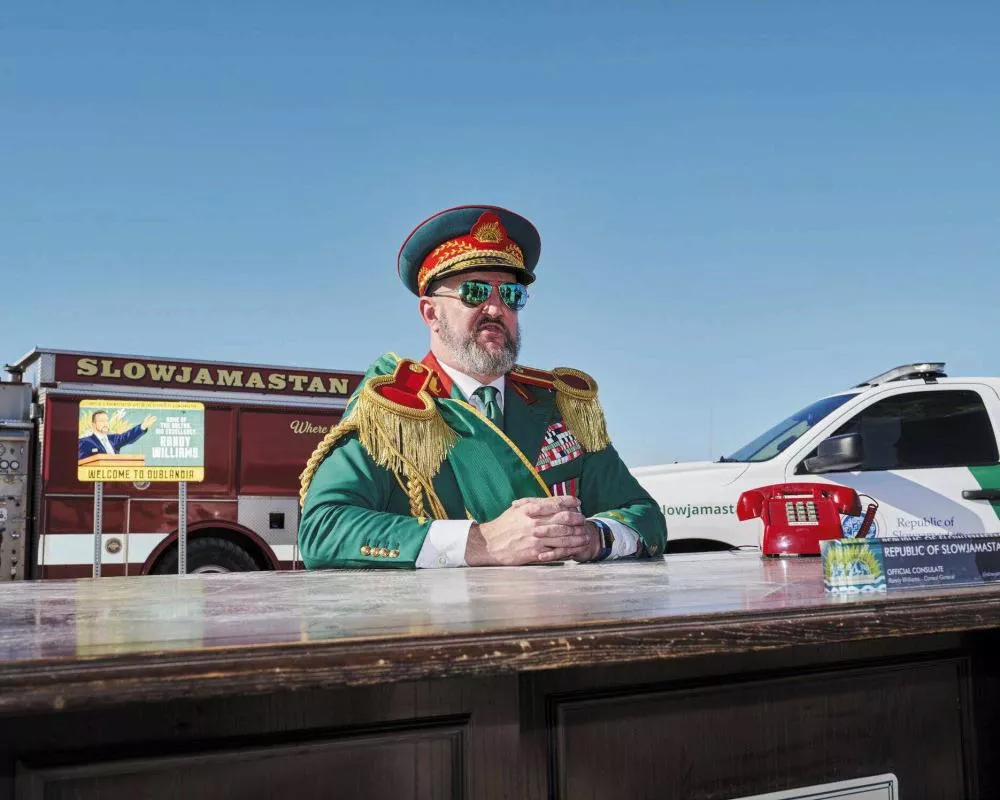 Un homme en uniforme militaire coloré se tient derrière un bureau en plein air. Derrière lui, on voit un camion avec l'inscription "SLOWJAMASTAN" et une voiture blanche. Une pancarte indique "Welcome to Dublania."