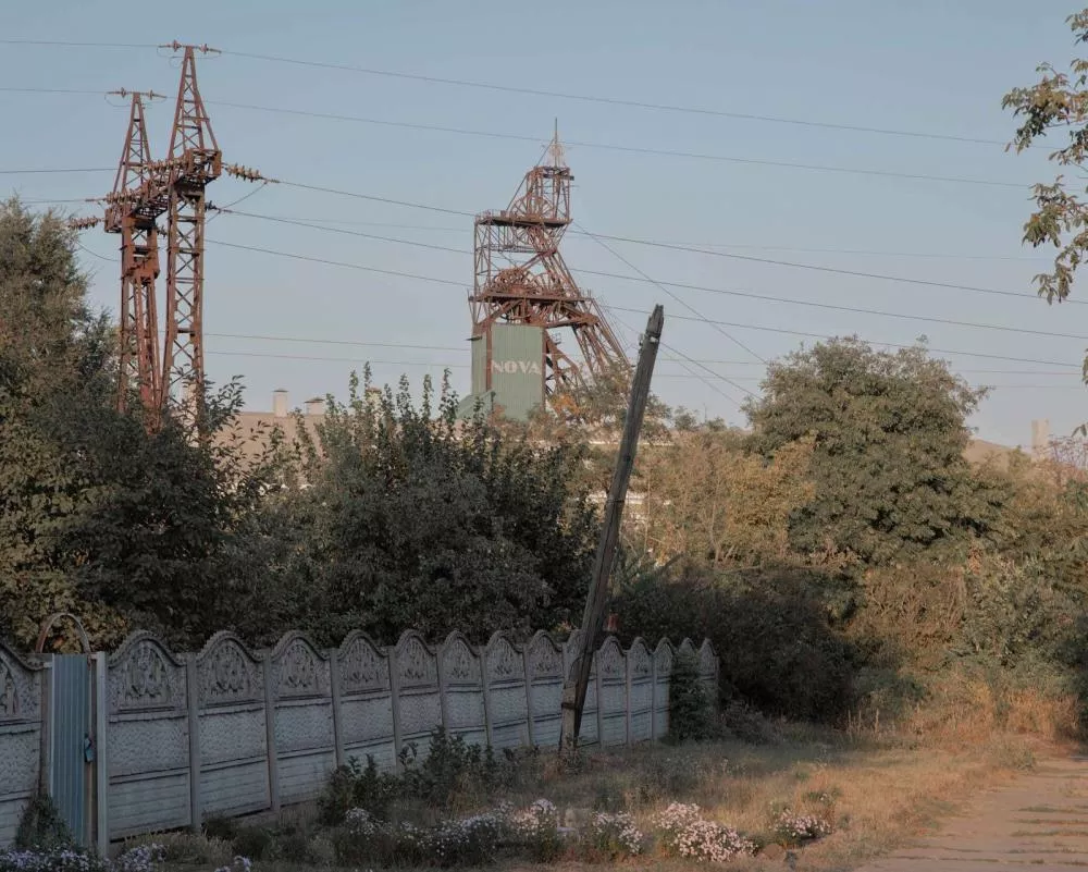 Une zone industrielle avec des structures métalliques de pylônes électriques et une tour d'extraction minière. Il y a des arbres et un chemin bordé d'une clôture décorative en béton.