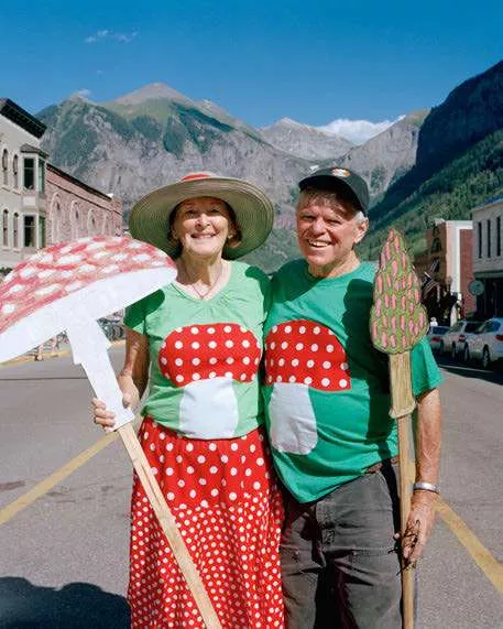 Un couple souriant, habillé de manière assortie avec des motifs de champignons rouges à pois blancs, se tient dans une rue avec des montagnes en arrière-plan. Ils tiennent chacun une décoration en forme de champignon.