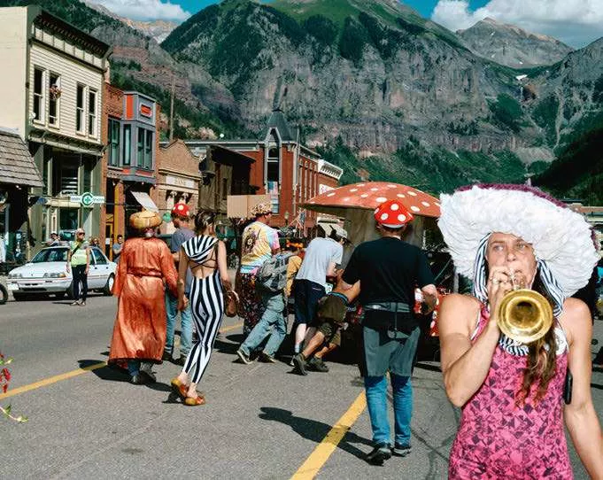 Une scène de rue animée dans une ville entourée de montagnes, avec des personnes costumées, dont certaines portent des chapeaux en forme de champignon. Une femme joue de la trompette au premier plan.