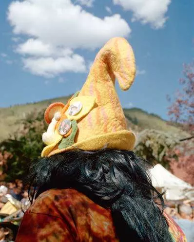 Une personne porte un chapeau pointu jaune orné de badges et de décorations, avec des cheveux longs et noirs. En arrière-plan, on voit un paysage de montagne sous un ciel bleu avec quelques nuages.