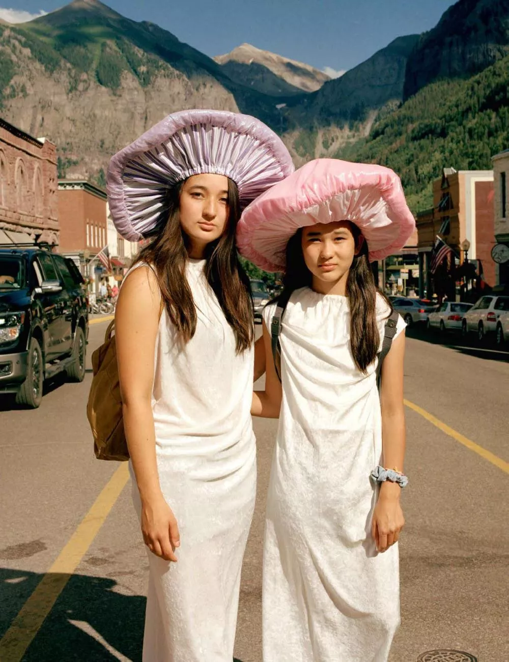Deux personnes vêtues de robes blanches et portant des chapeaux en forme de champignons se tiennent dans une rue avec des montagnes en arrière-plan.