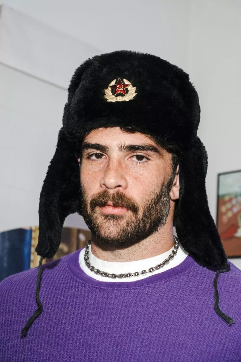 Un homme portant un chapeau de style russe avec un emblème soviétique, un pull violet et un collier en chaîne.