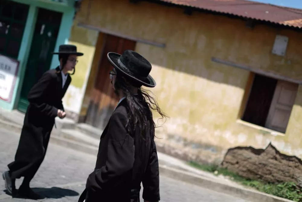Deux personnes portant des vêtements traditionnels sombres et des chapeaux noirs marchent dans une rue pavée devant un bâtiment à la façade jaune.