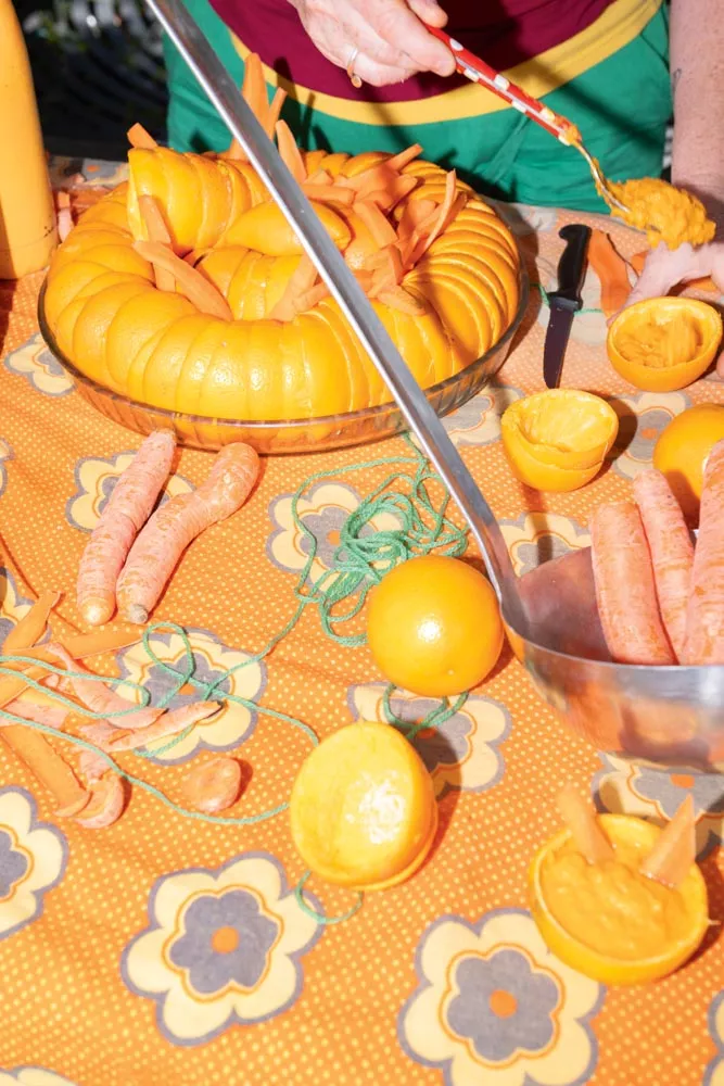 Une table est couverte d'une nappe orange à motifs floraux. Il y a des carottes, des oranges, une grande courge découpée, et une personne qui manipule les aliments.