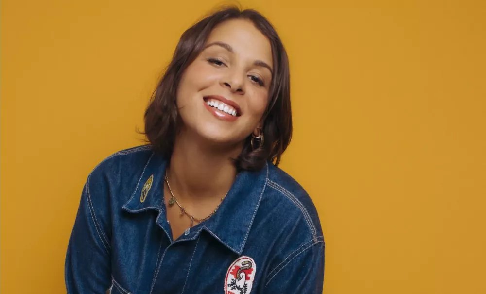 Une personne souriante portant une chemise en jean avec un fond jaune.