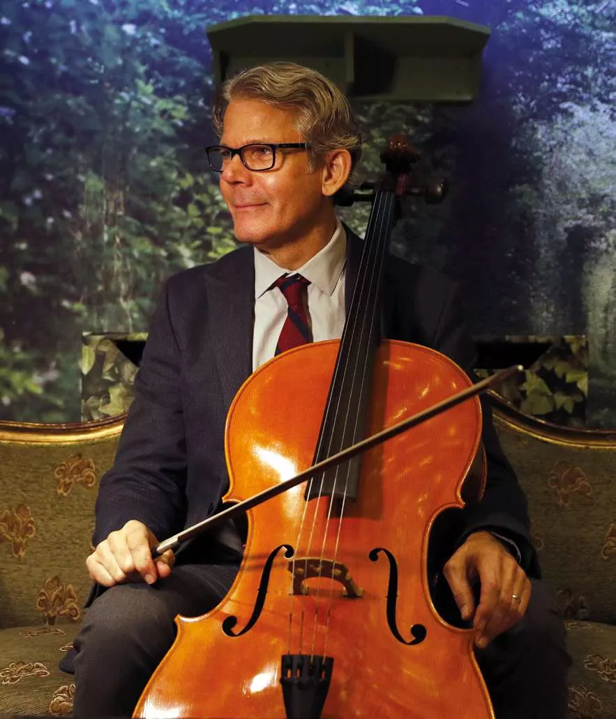 Un homme en costume et cravate est assis sur un canapé, tenant un violoncelle. Il porte des lunettes et semble être dans un environnement intérieur avec un fond de nature.