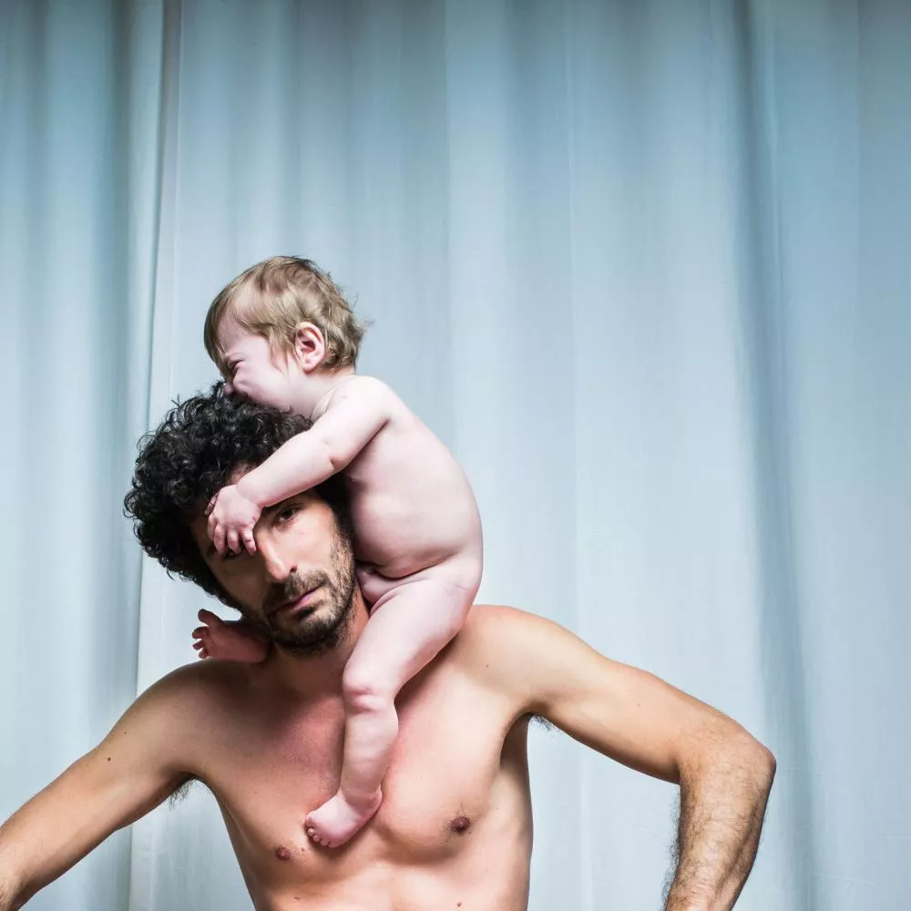 Un homme torse nu porte un bébé sur ses épaules. Le bébé est également nu et semble mordre ou jouer avec les cheveux de l'homme.