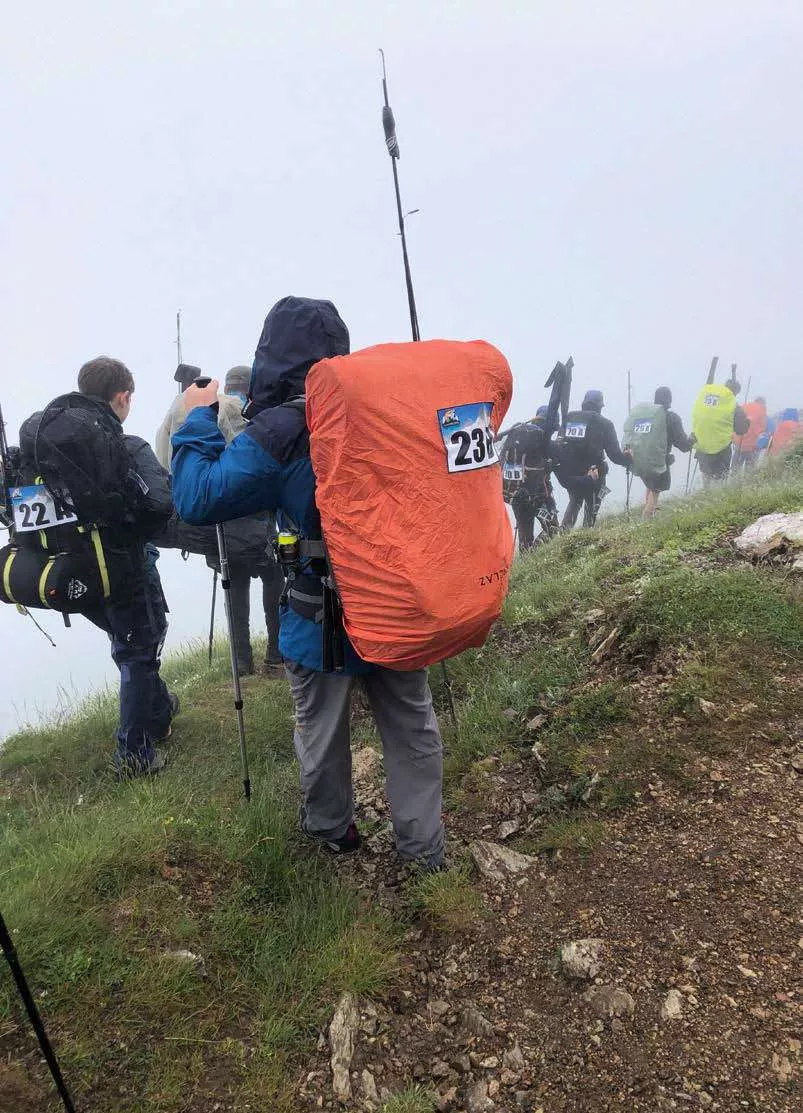 Des personnes participent à une randonnée ou une compétition en montagne, équipées de sacs à dos avec des housses imperméables et des bâtons de marche. Les participants portent des dossards numérotés.