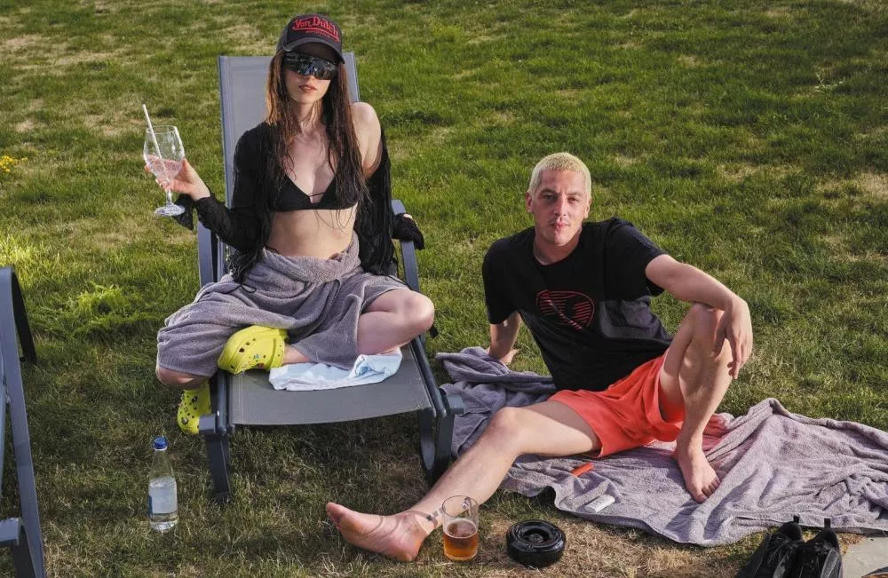 Deux personnes sont assises sur des chaises longues dans un jardin. La première personne porte un bikini noir, un chapeau et des lunettes de soleil, tenant un verre. La seconde personne porte un t-shirt noir et un short orange, assise sur une serviette avec un verre de bière à côté.