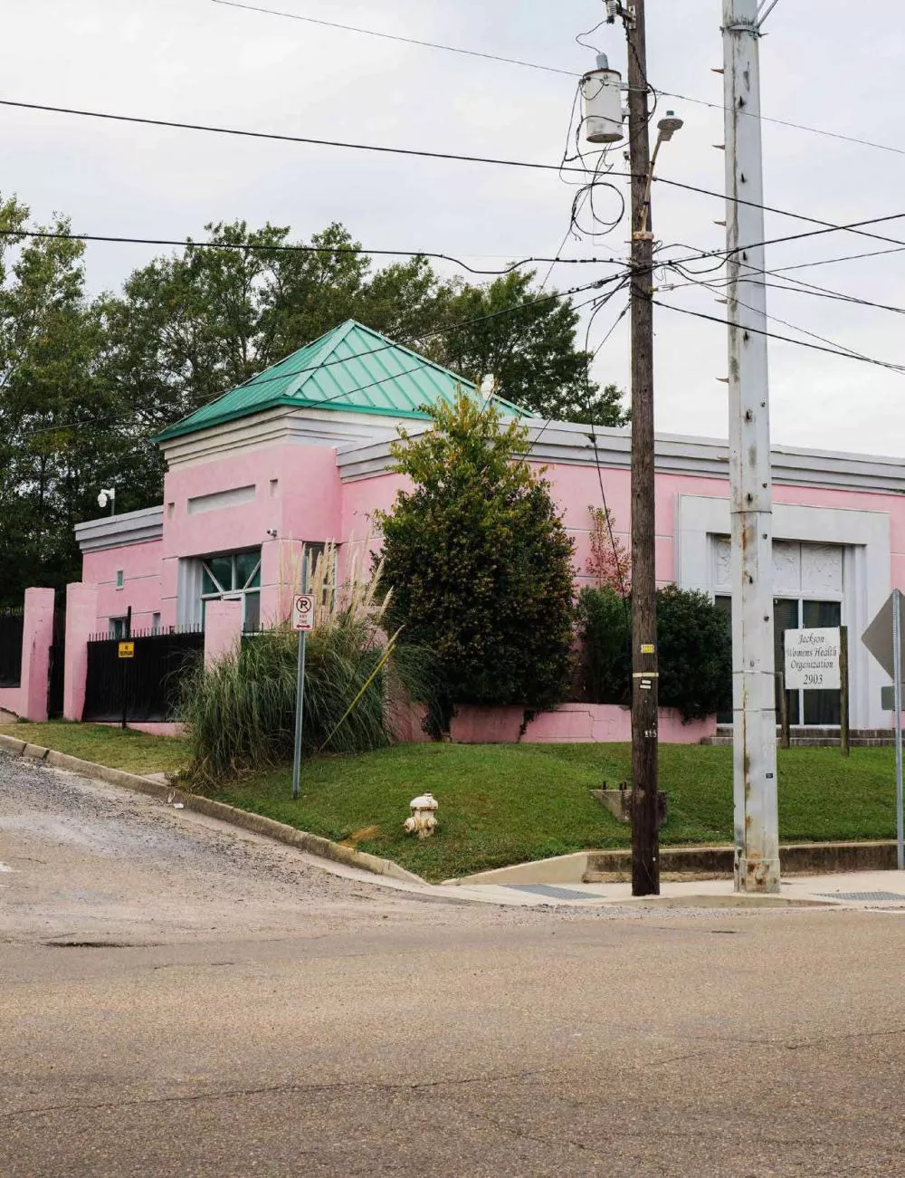 Un bâtiment rose avec un toit vert, entouré de verdure, se trouve à l'angle d'une rue. Il y a des poteaux électriques et des câbles au premier plan. Un panneau indique "Jackson Women's Health Organization 2903".
