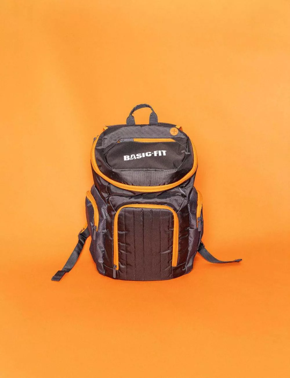 Un sac à dos noir avec des détails orange portant l'inscription "BASIC-FIT" sur un fond orange.