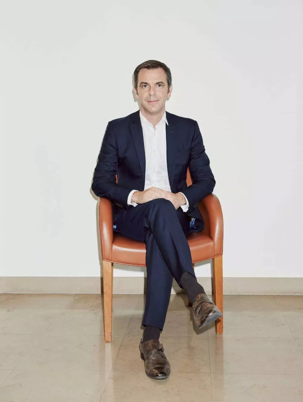Un homme en costume sombre est assis sur une chaise en cuir marron, les jambes croisées, dans un environnement sobre avec un sol en marbre clair.