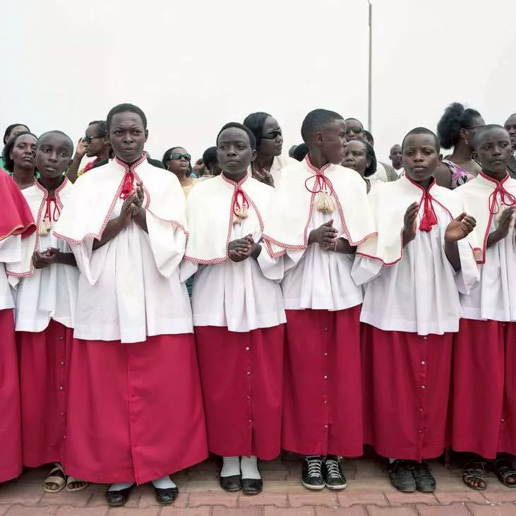 Un groupe de jeunes vêtus de tenues liturgiques rouges et blanches se tient debout, les mains jointes ou en train d'applaudir, entouré de plusieurs adultes en arrière-plan.
