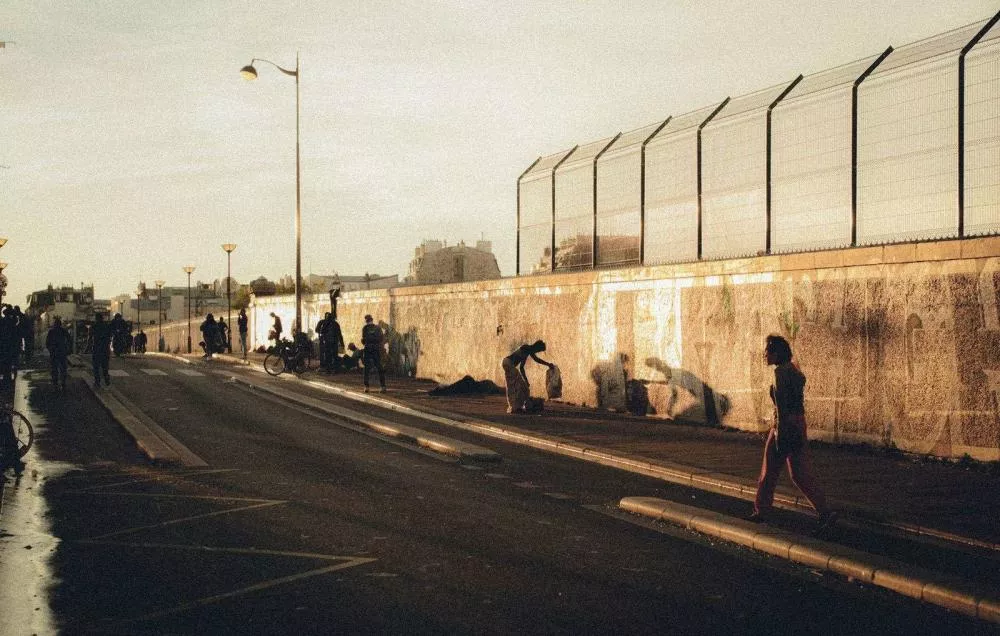 La photo montre une rue avec des personnes marchant le long d'un trottoir. Un mur orné de graffitis est visible à droite, et le soleil bas projette de longues ombres. L'atmosphère est calme avec une lumière dorée.
