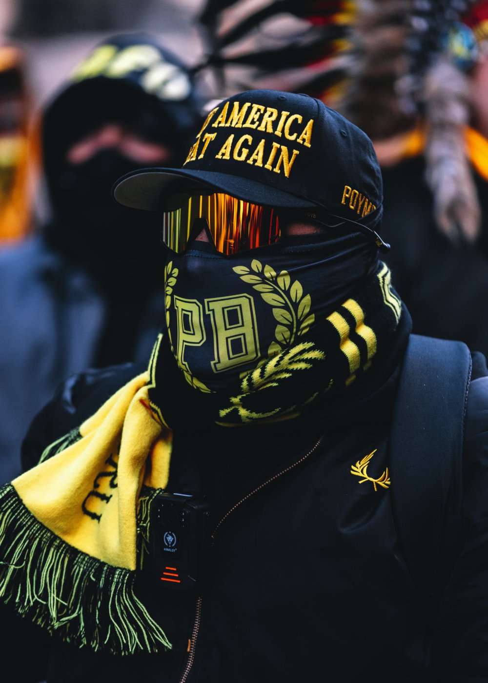 Un individu porte une casquette avec l'inscription "America Great Again", un masque avec les lettres "PB", des lunettes de soleil réfléchissantes, une écharpe jaune et noire, et une veste noire avec un emblème doré.