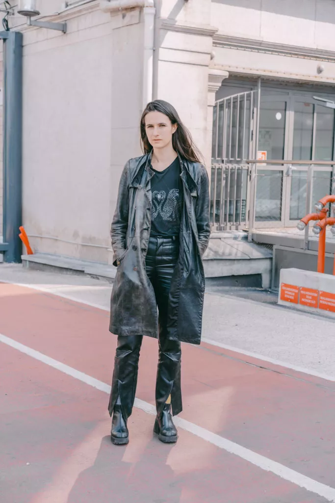 Une personne portant un long manteau en cuir noir, un t-shirt noir avec un motif, un pantalon en cuir noir et des bottes noires, se tient debout sur une surface rouge près d'un bâtiment.