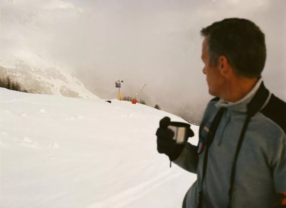 Un homme en tenue de ski tient une tasse dans la main, regardant vers un paysage enneigé avec des panneaux de signalisation sur une piste de ski.