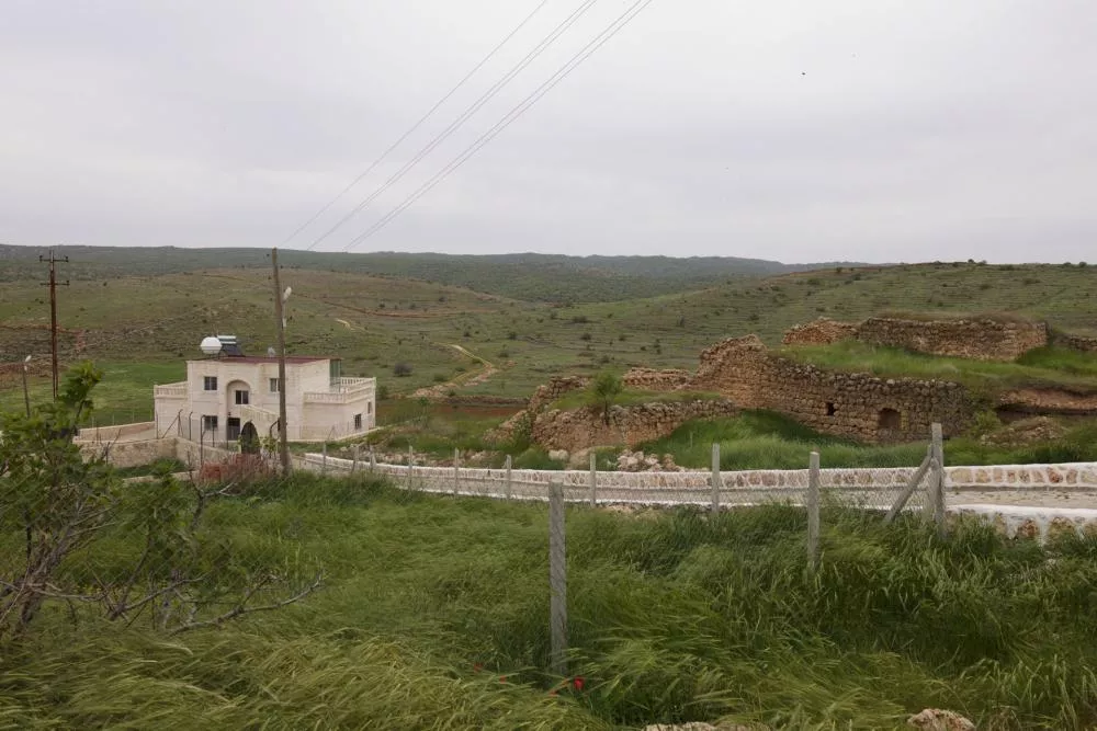 Une maison blanche moderne se dresse à côté de ruines en pierre dans un paysage rural verdoyant, avec des collines en arrière-plan. Un chemin pavé et une clôture en grillage entourent la propriété.