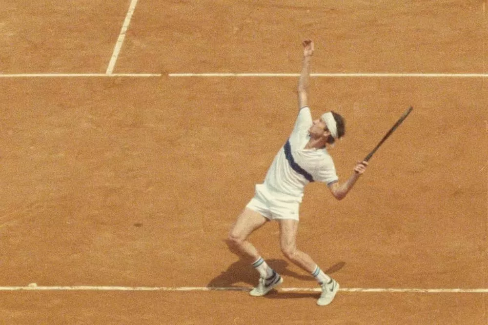 Un joueur de tennis est en train de servir sur un court en terre battue. Il porte un bandeau, un polo blanc avec une bande bleue, un short blanc et des chaussures de sport blanches.