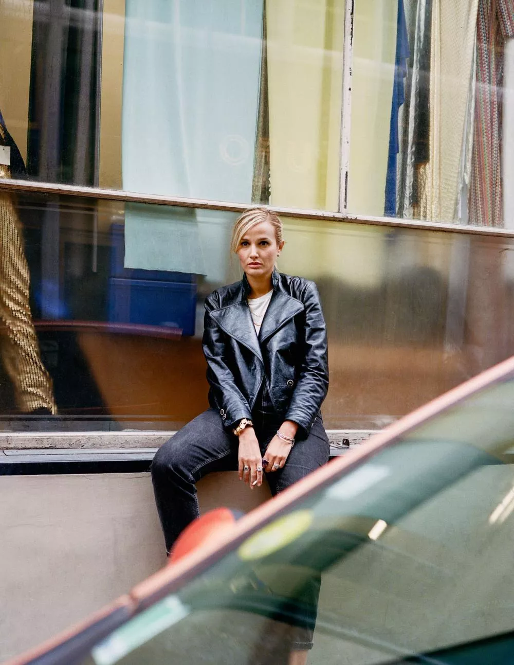 Une personne assise, portant une veste en cuir noir, posant devant une façade vitrée.