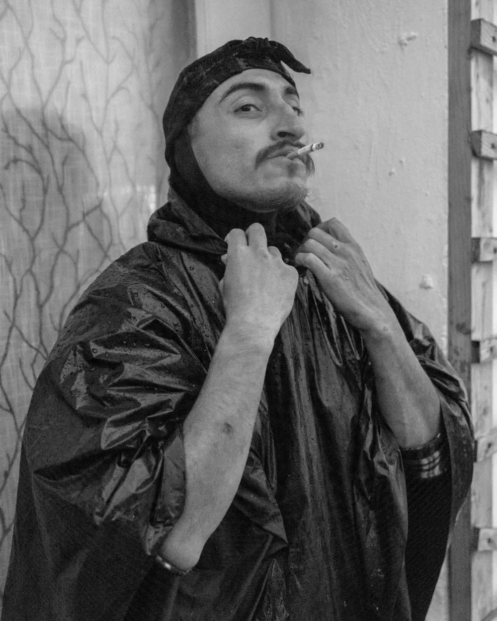 Un homme portant un poncho imperméable et un bonnet, tenant une cigarette à la bouche. Il semble être sous la pluie.