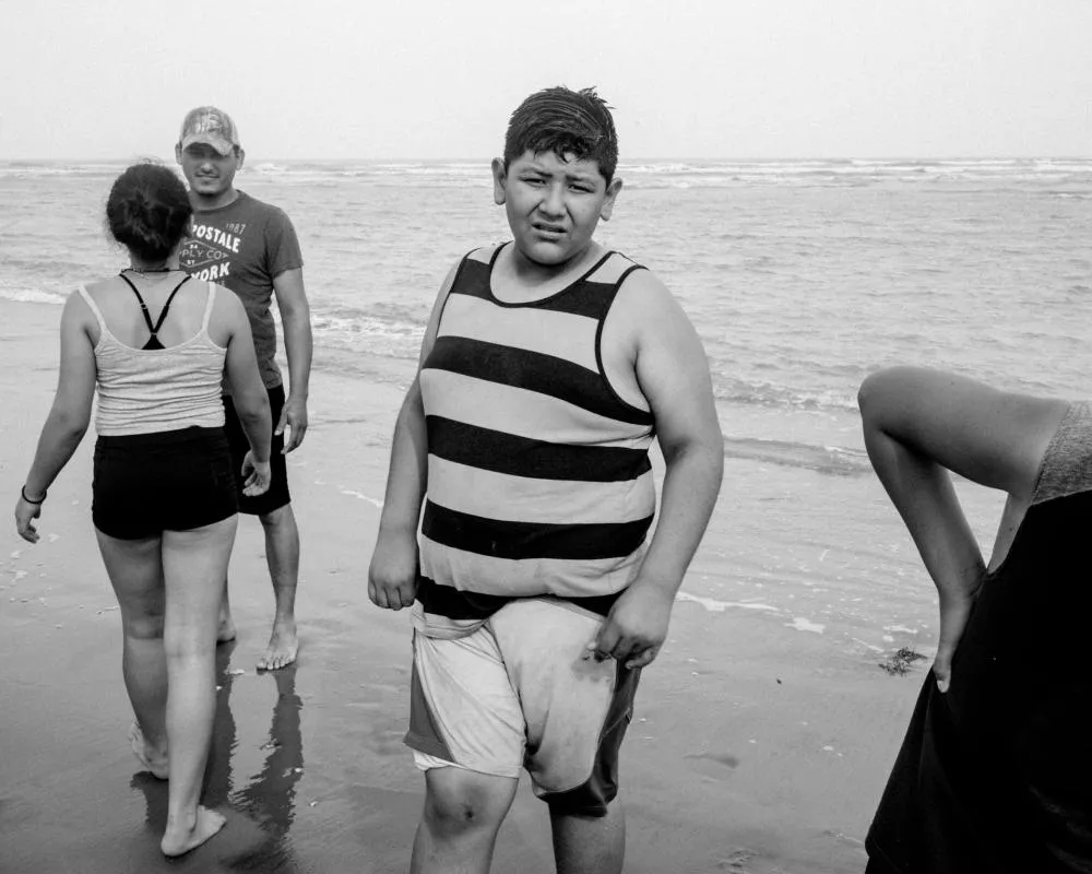 Une scène de plage en noir et blanc avec plusieurs personnes marchant sur le sable près de l'eau. Un garçon au centre porte un débardeur à rayures.