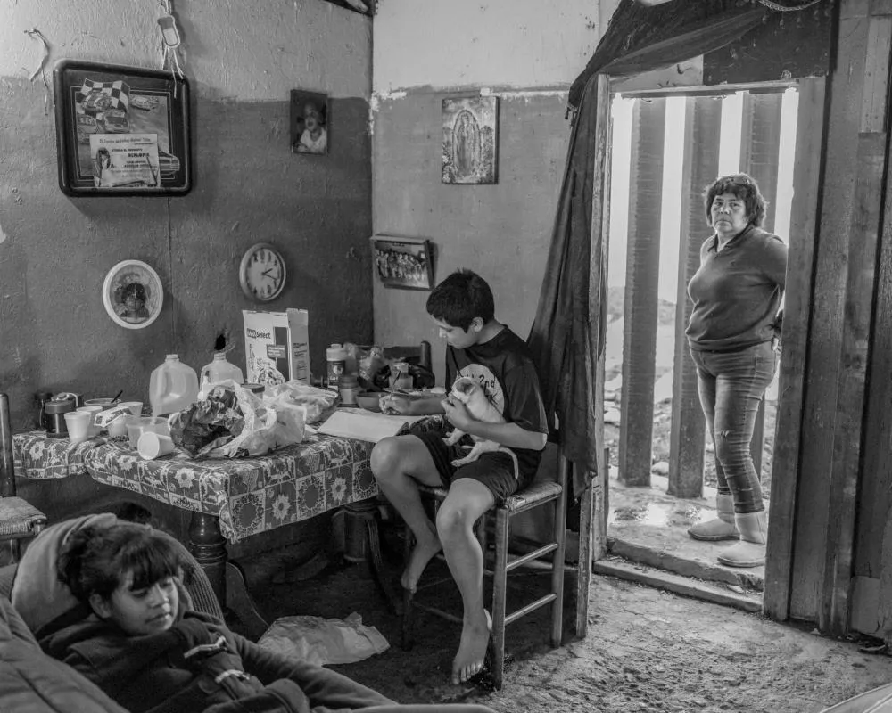 Une pièce simple avec des murs décorés de quelques images et une horloge. Un garçon est assis à une table avec des aliments et un carnet, tenant un petit chien. Une femme se tient à l'entrée, regardant à l'intérieur, tandis qu'une fille est assise sur un canapé.