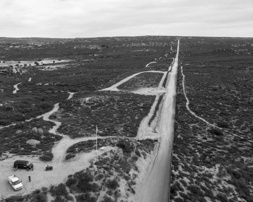 Une route droite traverse un paysage désertique avec des chemins de terre sinueux. Deux véhicules et un quad sont stationnés près d'un mât de drapeau.