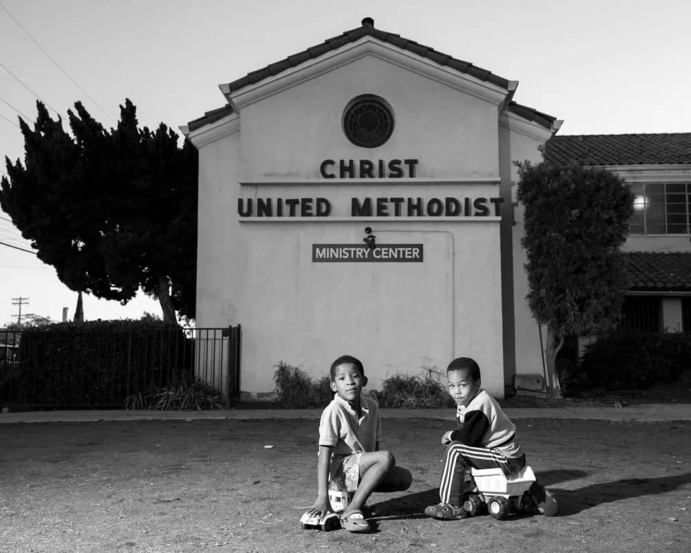 Deux enfants sont assis devant un bâtiment avec l'inscription "Christ United Methodist Ministry Center". L'un des enfants joue avec un camion jouet.