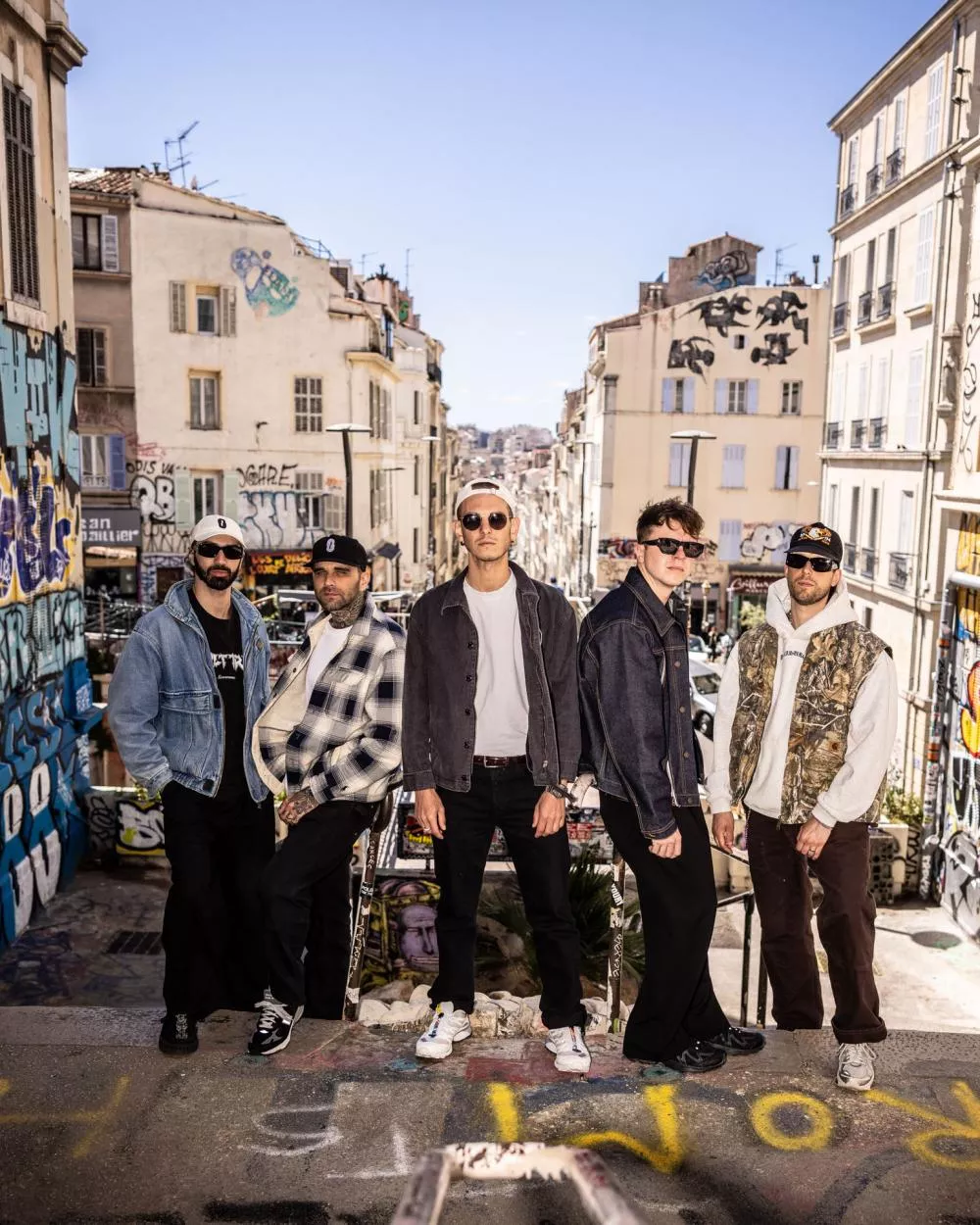 Cinq personnes posent dans une rue urbaine, entourées de bâtiments avec des graffitis. Ils portent des vêtements décontractés, comme des vestes en jean et des casquettes. L'arrière-plan montre un quartier avec des immeubles et des œuvres de street art.
