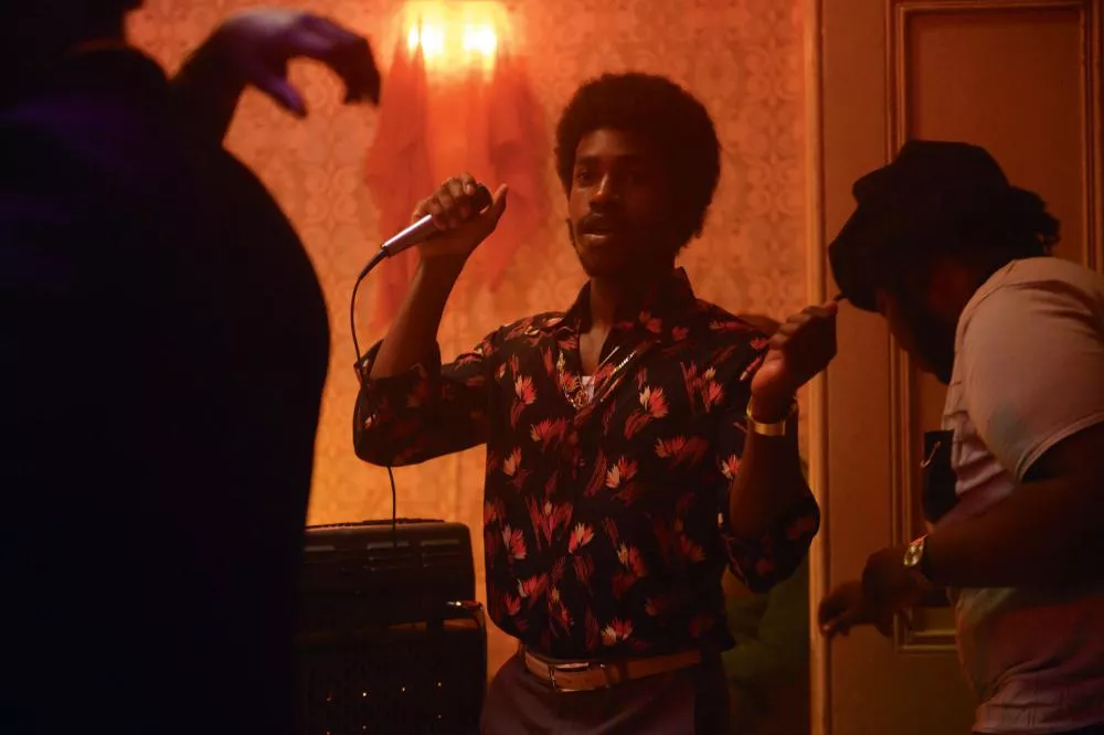 Un homme tenant un micro chante ou parle dans une ambiance tamisée, avec une chemise à motifs colorés. Une autre personne est à côté de lui, semblant danser ou bouger au rythme de la musique.
