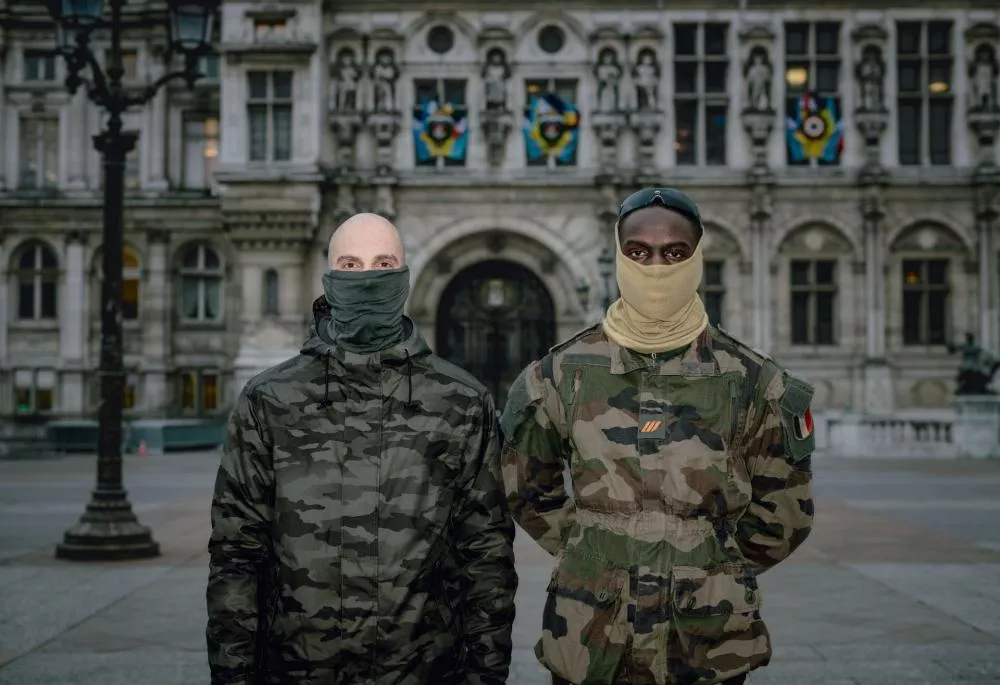 Deux personnes masquées portant des vêtements de camouflage se tiennent devant un bâtiment historique.