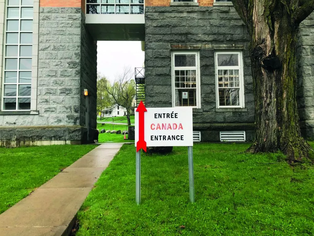 Un panneau indique "ENTRÉE CANADA ENTRANCE" avec une flèche rouge pointant vers le haut, situé devant un bâtiment en pierre grise avec des fenêtres.