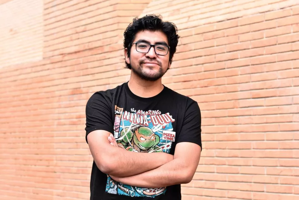 Un homme avec des lunettes et une barbe légère, portant un t-shirt noir avec un motif de tortue ninja, se tient les bras croisés devant un mur de briques.