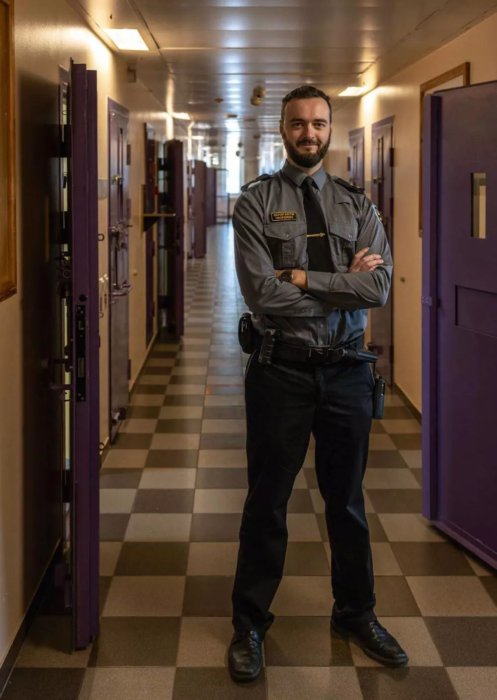 Un homme en uniforme de gardien de prison se tient dans un couloir avec des portes de cellules violettes.
