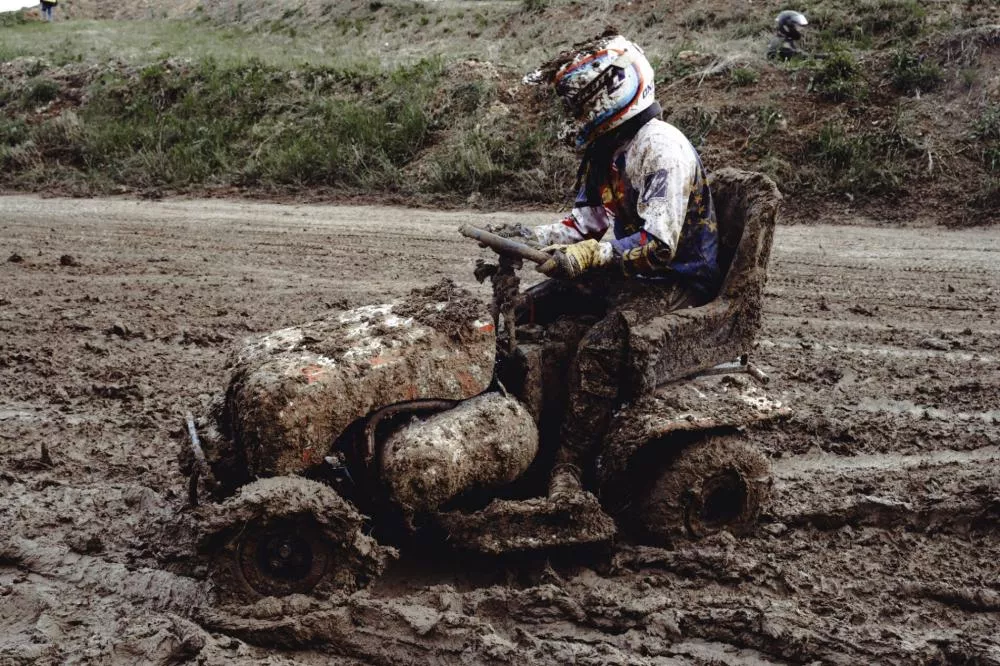 Un pilote en tenue de protection et casque conduit une tondeuse à gazon couverte de boue sur un terrain boueux.