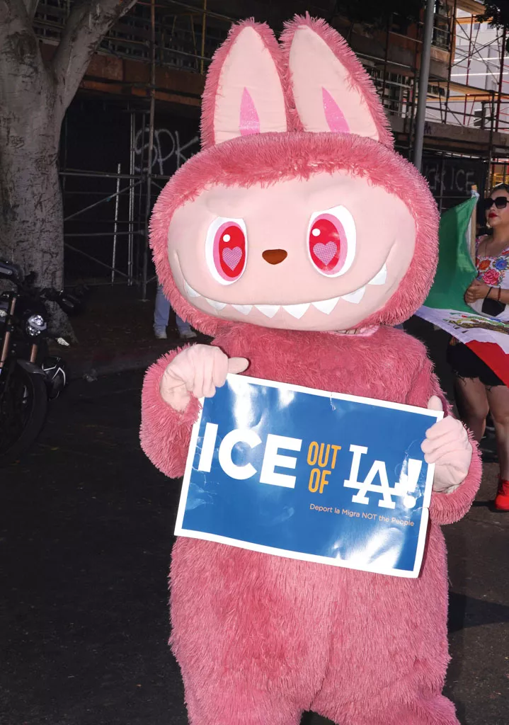 Un personnage en costume rose avec des oreilles de lapin tient une pancarte disant "ICE OUT OF LA! Deport la Migra NOT the People".