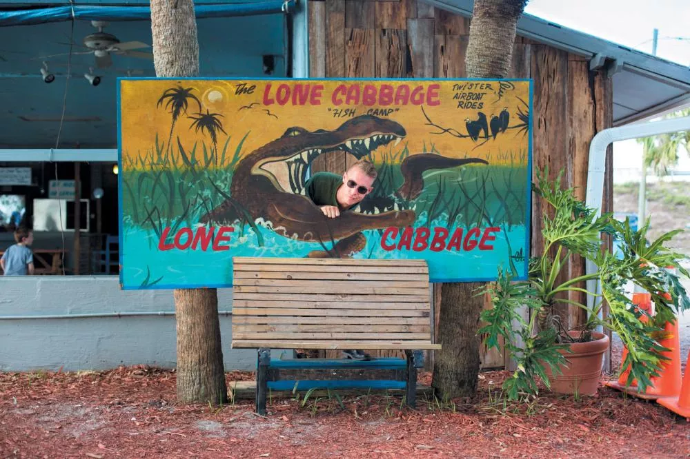 Un homme portant des lunettes de soleil pose derrière un panneau coloré représentant un crocodile, avec l'inscription "The Lone Cabbage Fish Camp".