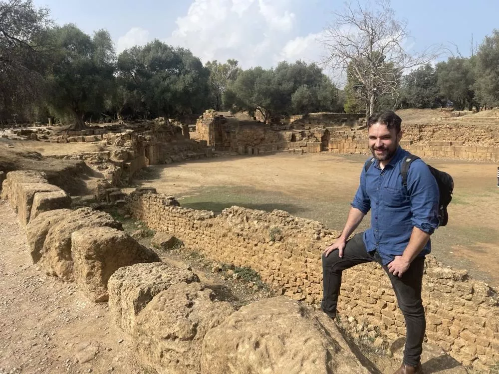 Un homme portant une chemise bleue et un sac à dos pose devant des ruines anciennes entourées d'arbres.