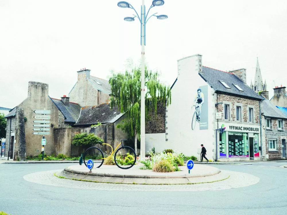 Une rue avec un rond-point où se trouve une sculpture de vélo. En arrière-plan, des bâtiments en pierre, dont un avec une peinture murale représentant un cycliste. Une agence immobilière est visible avec des panneaux de signalisation indiquant des directions.