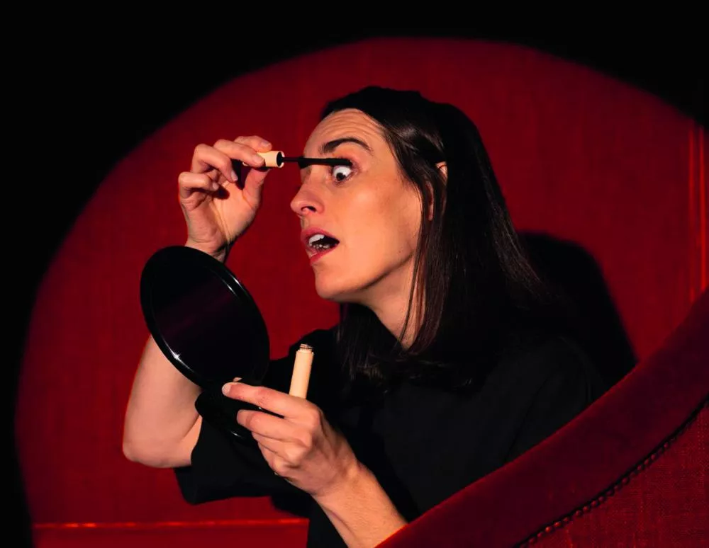 Une personne applique du mascara devant un miroir à main, assise sur un fauteuil rouge.