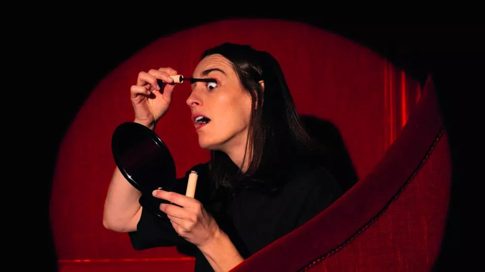 Une personne applique du mascara devant un miroir à main, assise sur un fauteuil rouge.