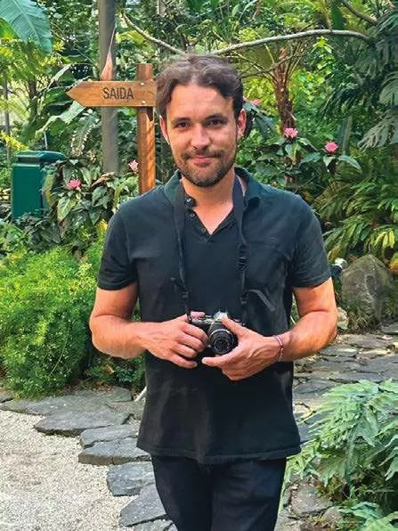 Un homme portant une chemise noire tient un appareil photo dans un jardin luxuriant. Un panneau indiquant "SAIDA" est visible en arrière-plan.