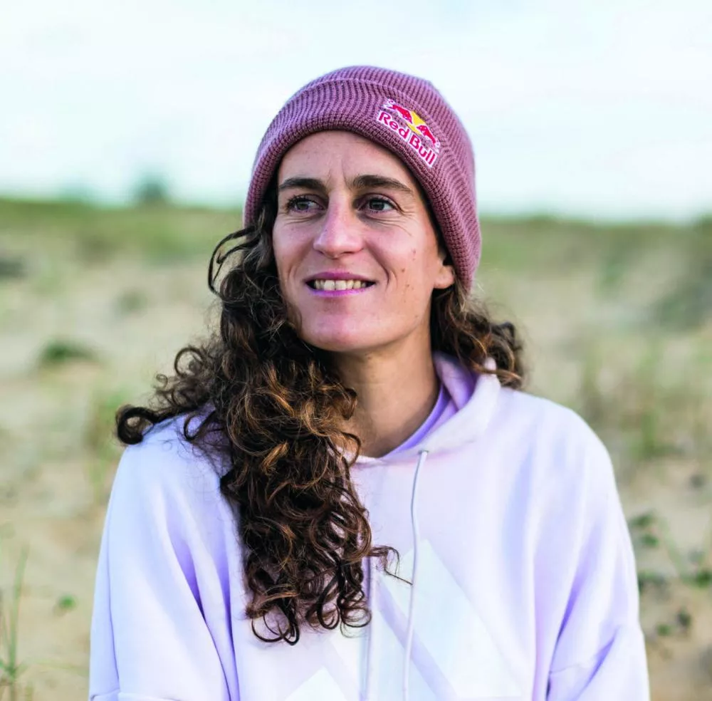 Une personne portant un bonnet rose avec un logo, un sweat à capuche blanc, assise sur du sable avec un arrière-plan de dunes.