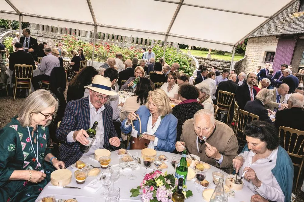 Un groupe de personnes est assis autour d'une table lors d'un événement en plein air sous une tente. Ils semblent être en train de déguster des plats et de discuter. L'ambiance est conviviale et festive.