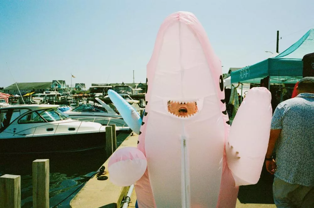 Une personne porte un costume gonflable de requin rose sur un quai avec des bateaux en arrière-plan.